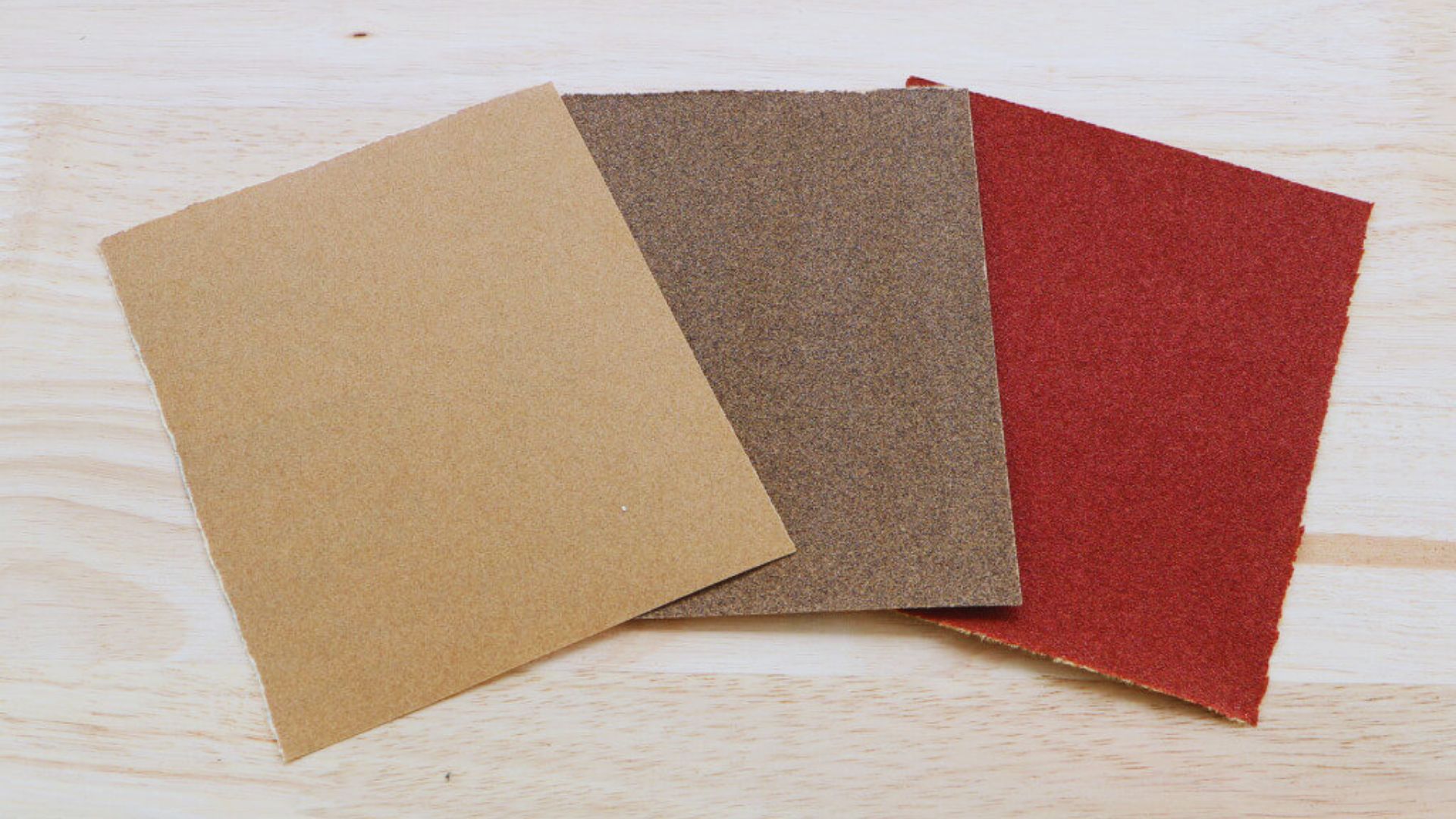 Three colored pieces of grit sandpaper for cabinets arranged on a table, showcasing different grits and textures.